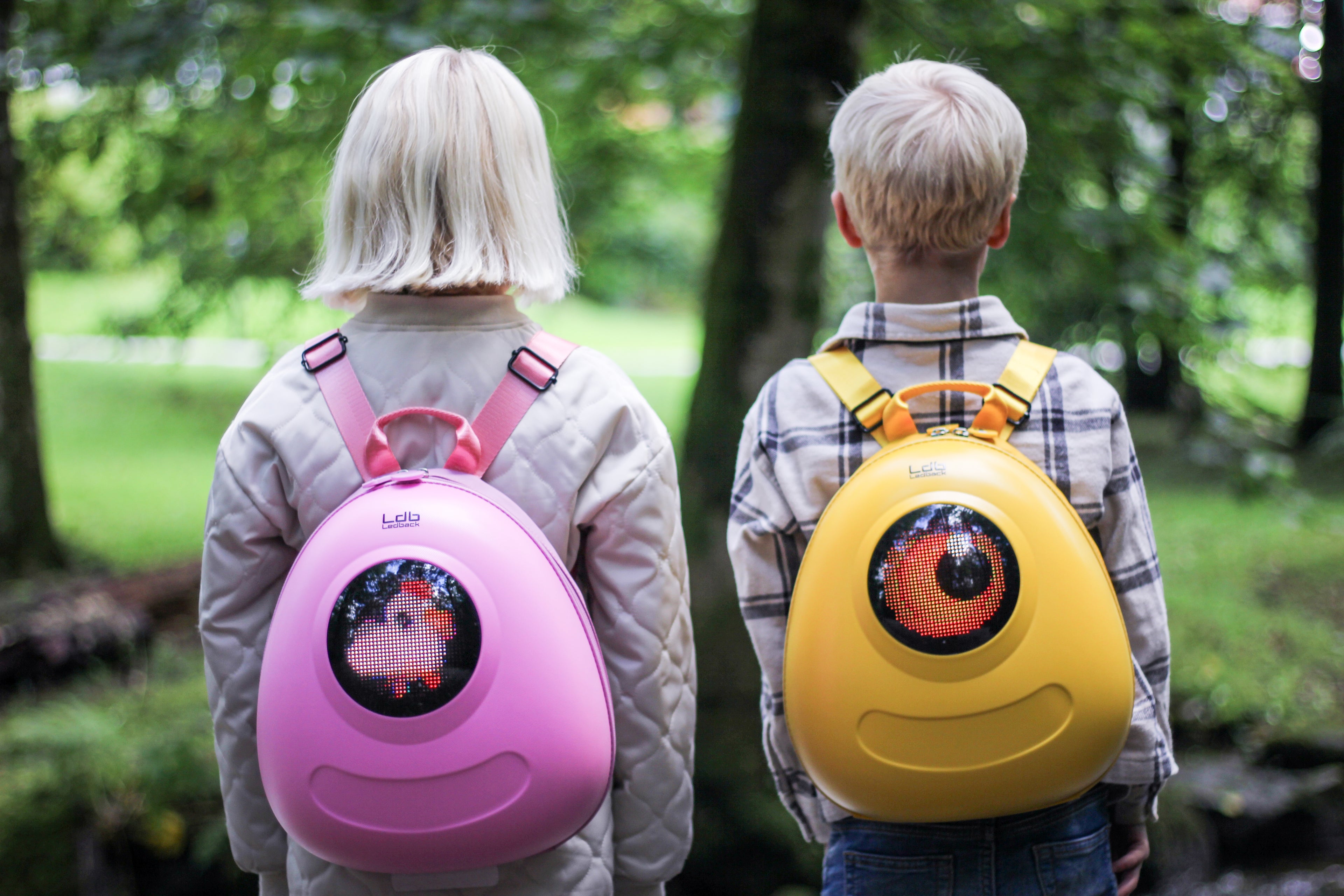 A girl and a boy showing off their Ledback Buddy backpacks. Girl with a Raspberry Sorbet Pink backpack with a white chicken on the round  LED screen, the boy with a Lemon Drop Yellow Buddy with an orange eye design on the LED screen.