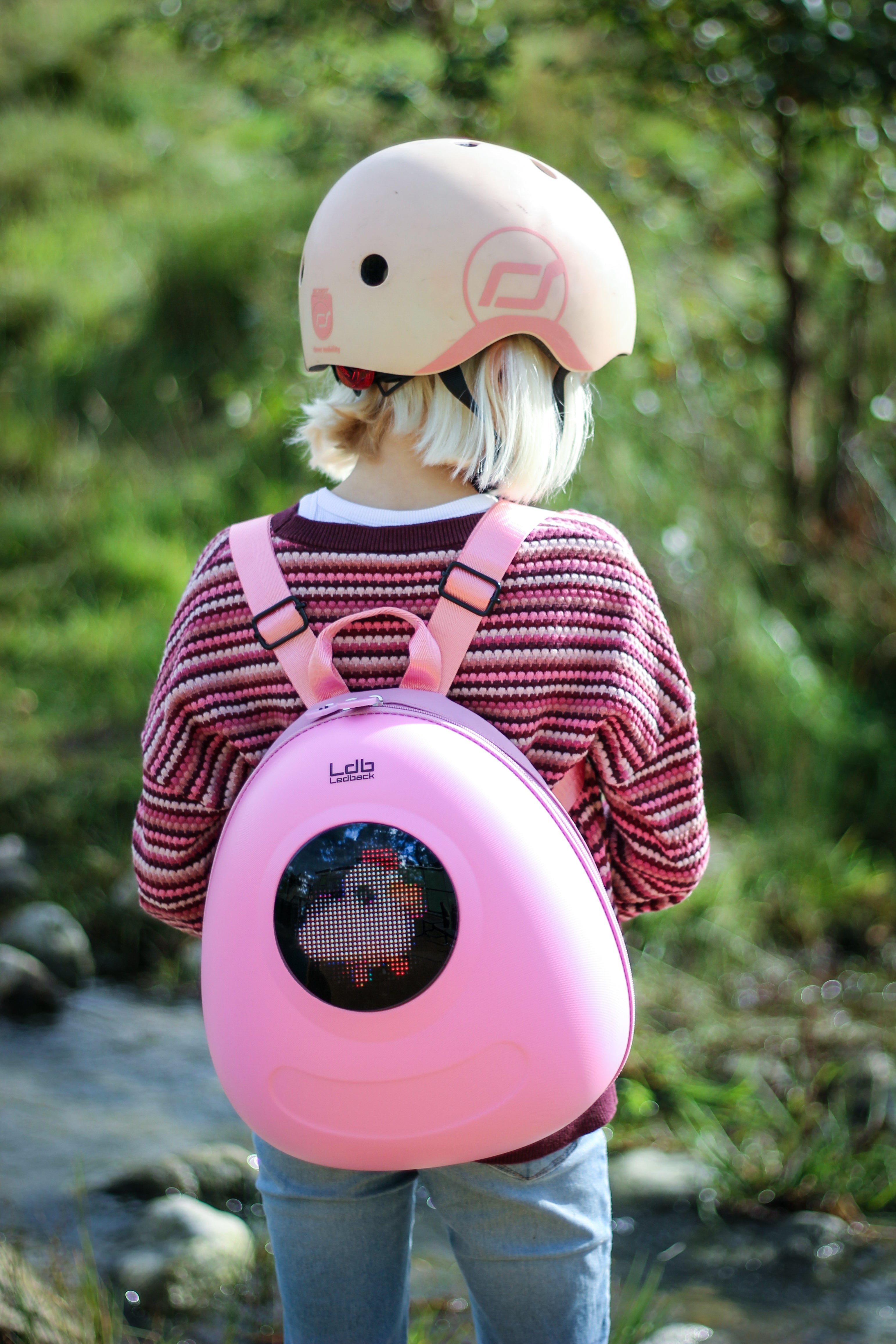 Girl on roller skates with a pink sweater and pink helmet, standing besides a stream in a playground, wearing a Raspberry Sorbet Pink Ledback Buddy backpack over her right shoulder, featuring a round LED screen. 