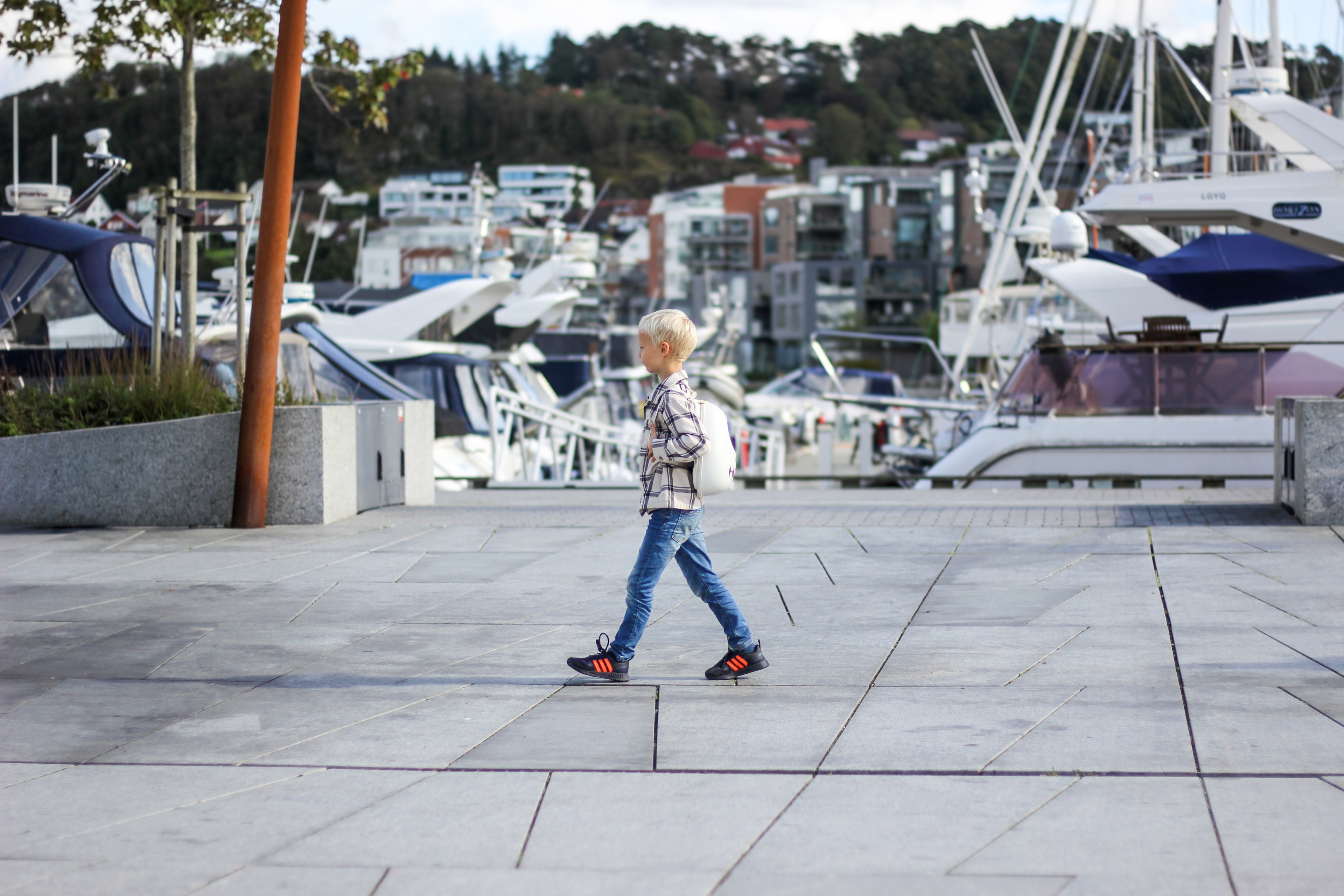 young boy "on a mission", walking straight across the camera view in the harbor in Sandnes, with boats in the background, wearing a Whipped Cream White Ledback Buddy backpack.