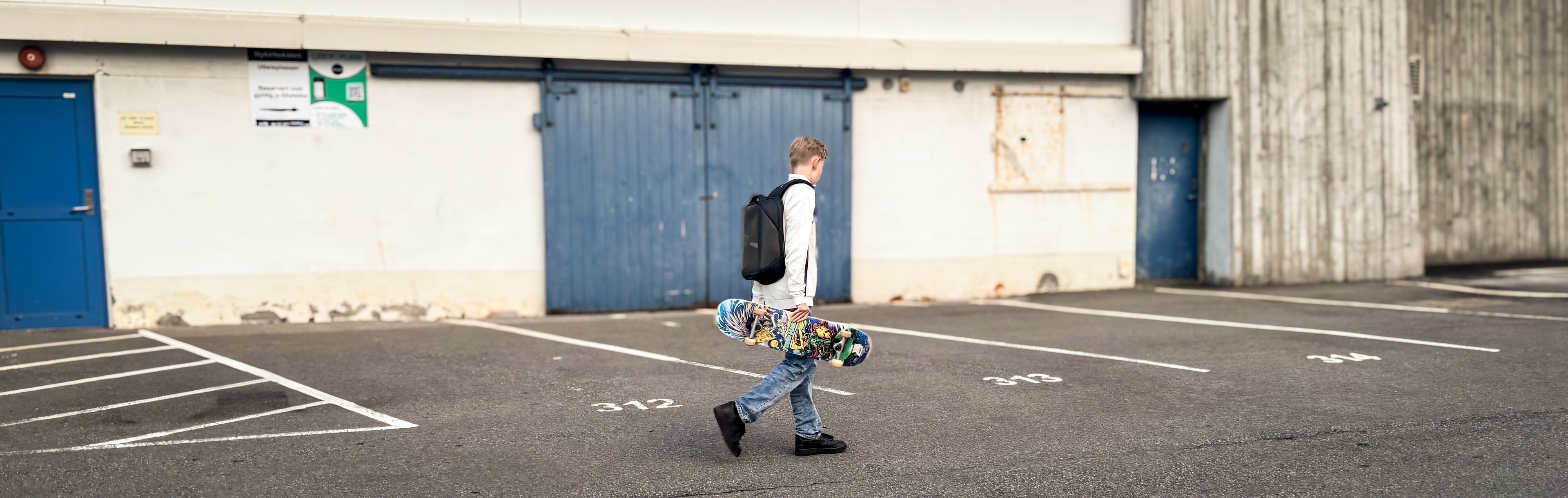 Adolescent boy walking with a colorful skateboard along storage houses in Bergen, wearing a Black Ldb Ledback HD Smart LED Laptop Backpack with the LED screen displaying a football game